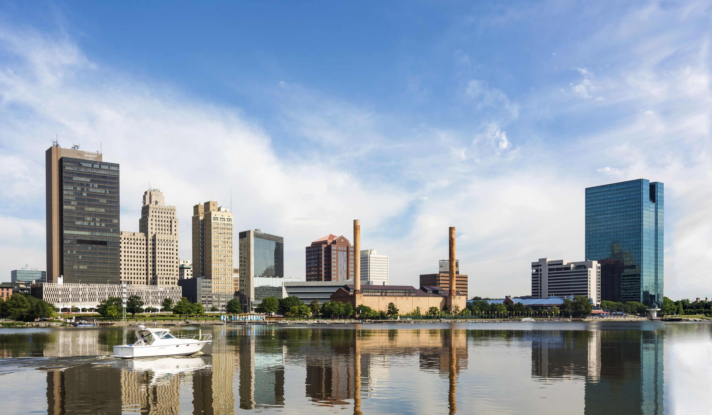 A panoramic view of downtown Toledo Ohio s skyline reflecting into the Maumee river with a power boat cruising by. A beautiful blue sky with white clouds for a backdrop.