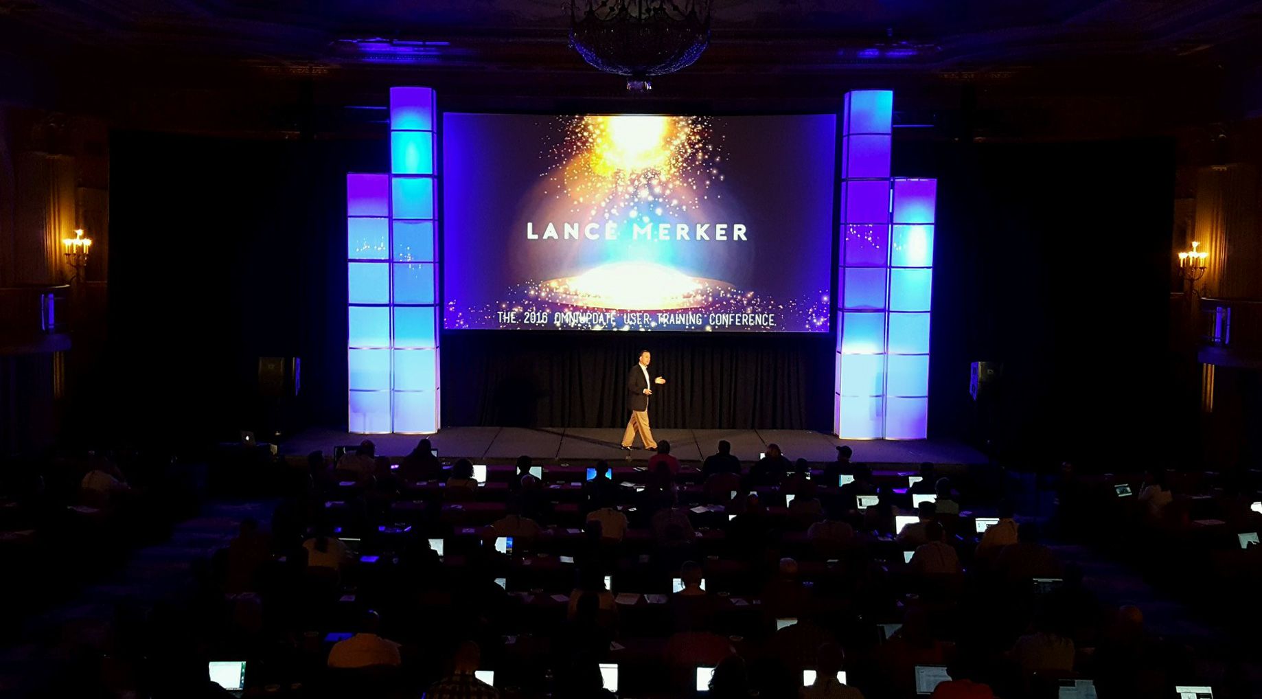 A man speaking on a conference stage with a large projection screen and plexiglass columns lit up with blue lighting