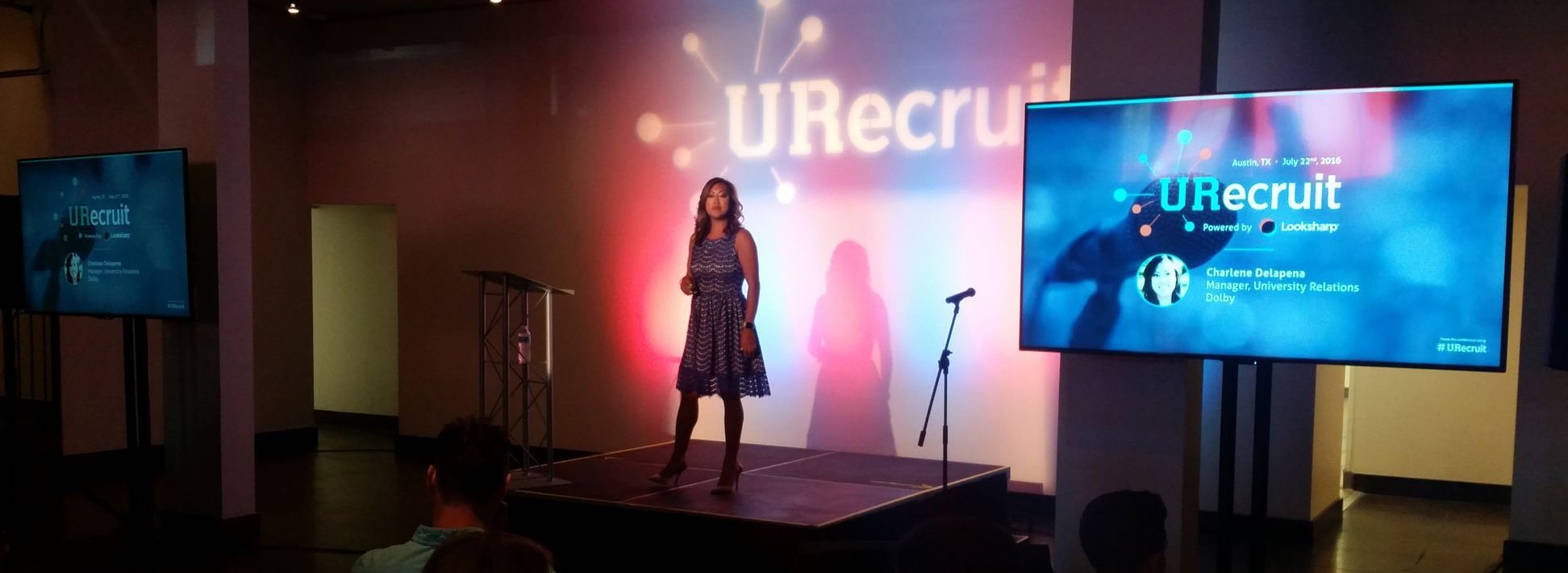 A woman speaking on a small stage with a two flat-screen TV display 