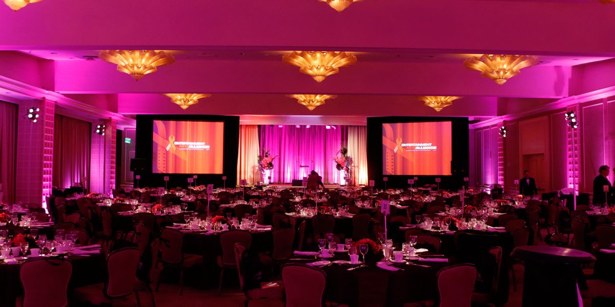 A large gala set up in a hotel ballroom with pink lighting and a two screen display 