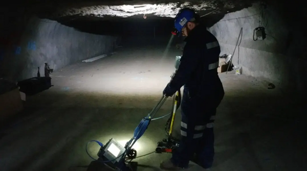 A man stands in left profile to the camera, with only the headlamp from his hardhat for light. The mine shaft he stands in quickly darkens beyond his light. He’s dressed in blue safety overalls with reflective tape, and is using ground penetrating radar equipment to scan the concrete slab at his feet.