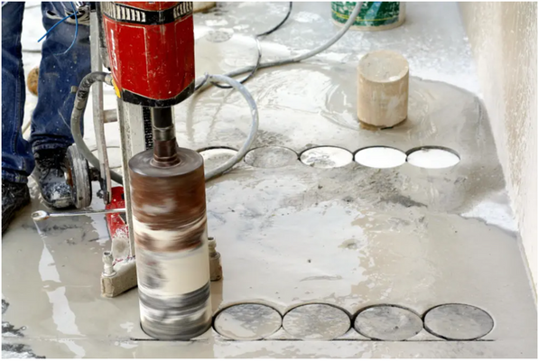 A red and black concrete coring machine being used on a wet concrete surface. Nine other holes have been drilled next to and behind the coring machine operated by a worker in jeans and black shoes. Concrete Coring Performed After GPRS Concrete Scanning