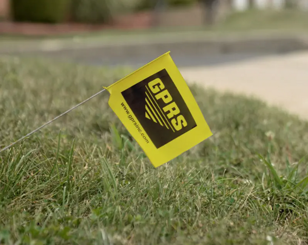 A yellow utility flag carrying GPRS’ logo in black marks the location of a gas line rises on a wire from a grassy area.