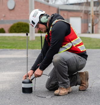 A GPRS Project Manager wearing a white helmet, green noise-cancelling headphones, a black hoodie, grey pants, a red vest, and brown shoes setting up an acoustic leak detector on asphalt.