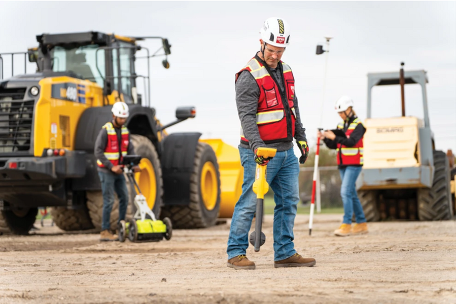 Three GPRS Project Managers, all dressed in boots, jeans, and safety gear, utilize an electromagnetic (EM) locator, a ground penetrating radar (GPR) cart, and a GNSS geolocating device at a construction site with heavy equipment in the background. 