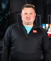 GPRS Project Manager Andrew Heine: A stocky white man with brown hair, dressed in a black GPRS quarter zip shirt over an aqua tee shirt, standing in front of a dark background. GPRS Project Manager Andrew Heine helped provide important data for a general contractor without impeding busy international airport operations.