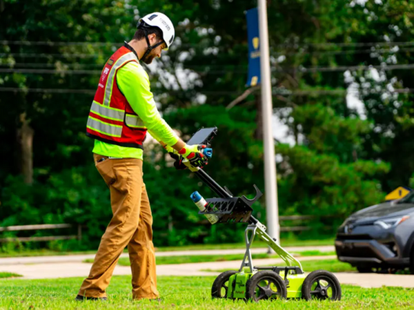 A GPRS Project Manager, glad in tan workpants, a reflective long-sleeved work shirt, a red safety vest, gloves, and a white GPRS helmet, employs a walk-behind ground penetrating radar scanner in a grassy area with sidewalks in the distance. A digital tablet is affixed to his cart, and there is a can of blue spray paint in a holder on the handles.