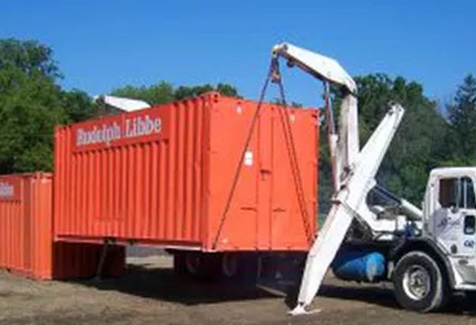 Orange conex box being lifted onto sidelifter trailer truck. 528-be-more-productive.jpg