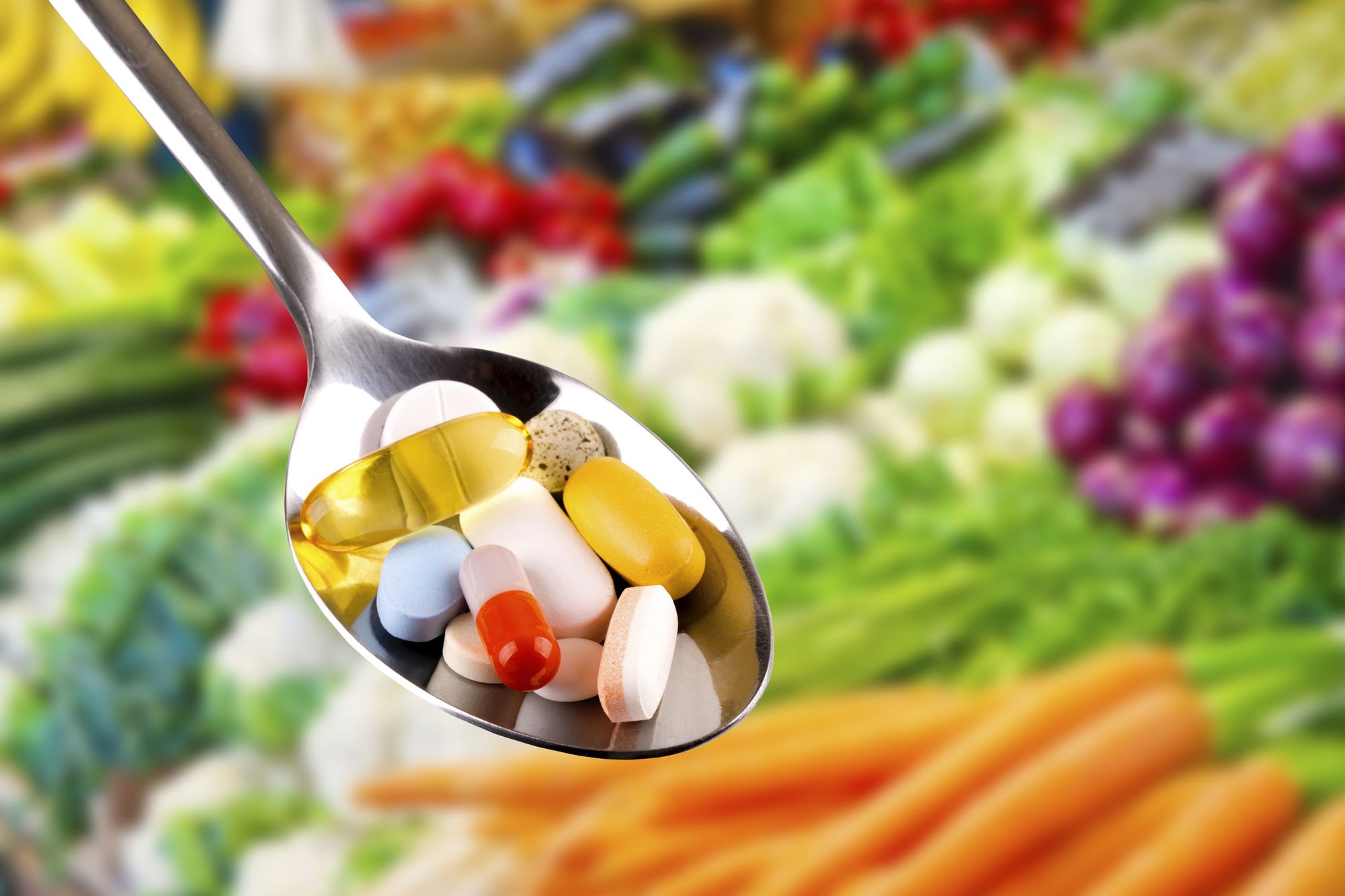 A spoon filled with assorted capsules and vitamins held over fresh vegetables. Spoonful of Supplements