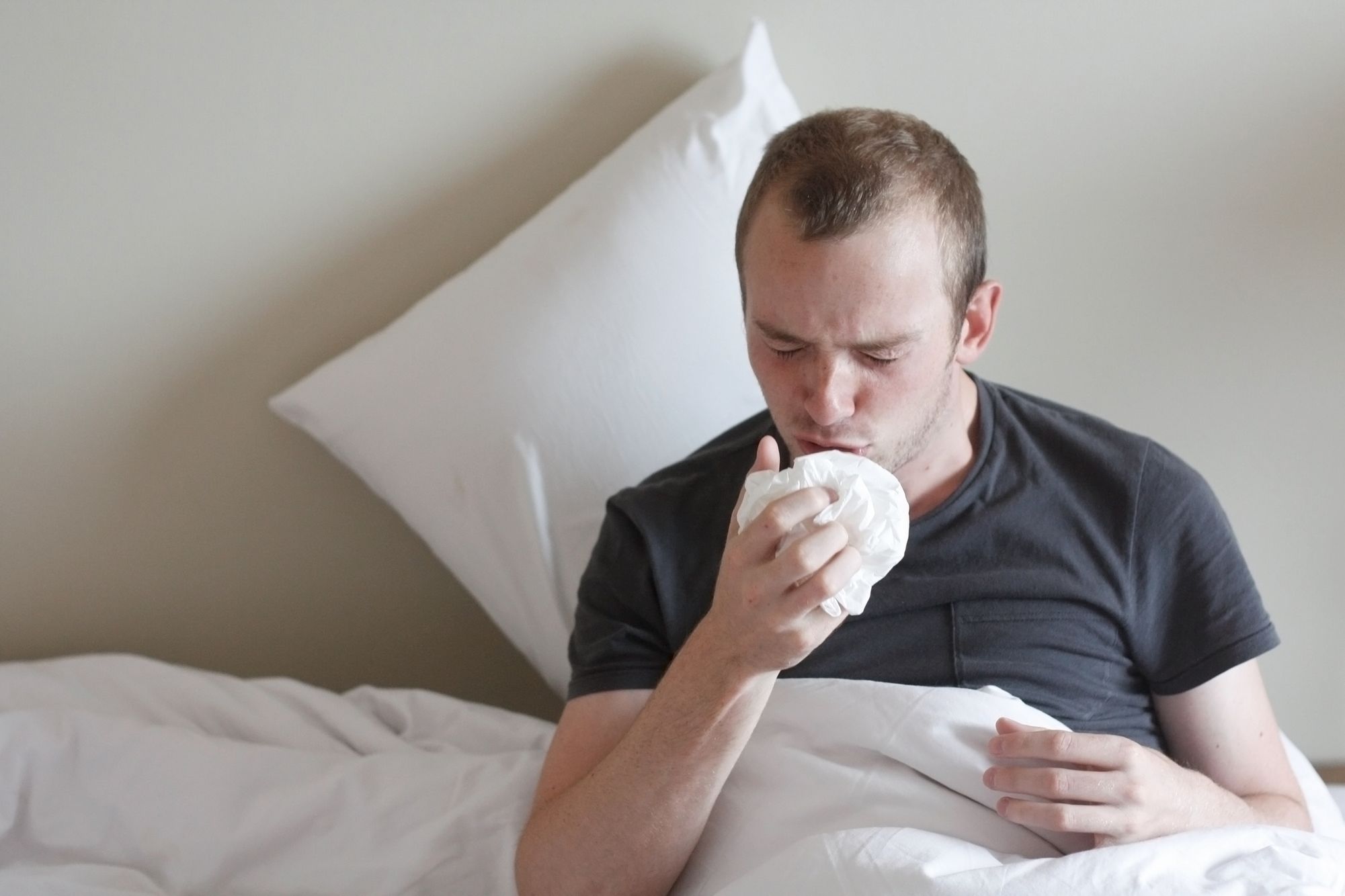 Man with short hair in a gray t-shirt sitting up in bed under the covers, sneezing into a tissue Sick Man In Bed