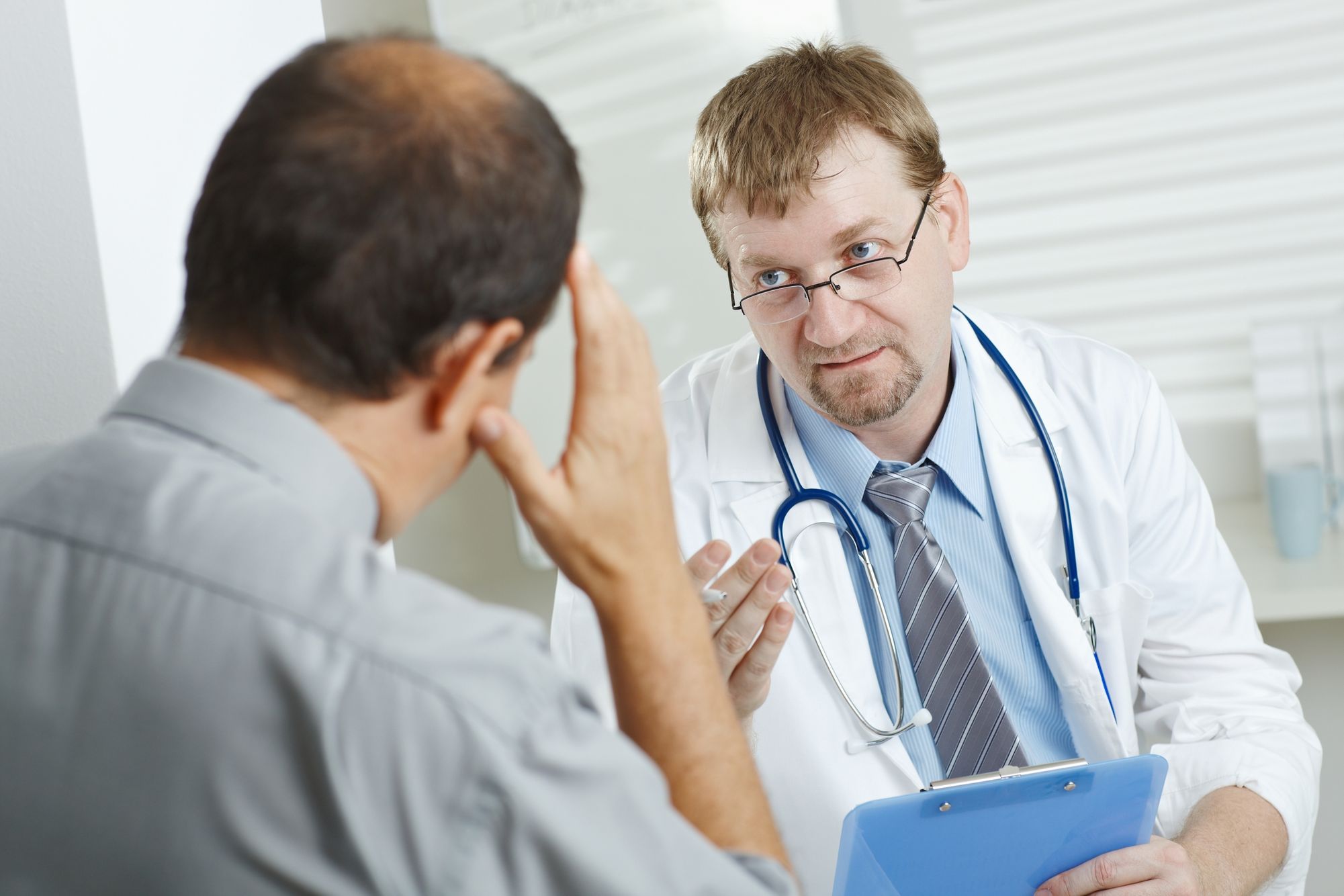Male doctor with glasses advising a patient that is facing away from the camera with his hand holding his head Doctor advising patient