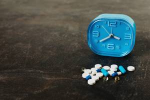 Blue alarm clock next to a pile of assorted prescription pills and capsules on a dark surface.
