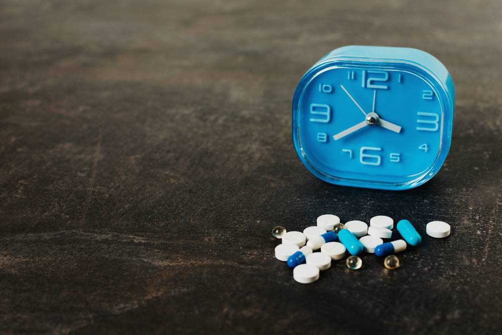 Blue alarm clock next to a pile of assorted prescription pills and capsules on a dark surface.