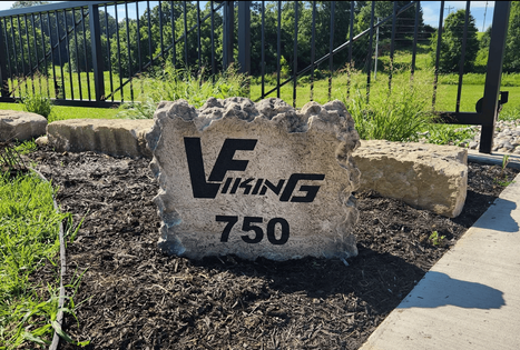 Commercial stone sign engraved with the “VF Viking” logo and the number 750, installed in landscaped mulch near a metal fence, created by Boulder Designs.