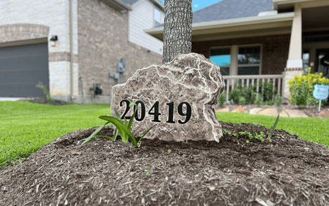 Residential landscape boulder engraved with the house number “20419,” installed in a mulch bed near a tree in a front yard, created by Boulder Designs.