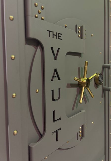 Steam punk finished grey vault door with gold rivets, a gold handle, and The Vault engraved in the front.