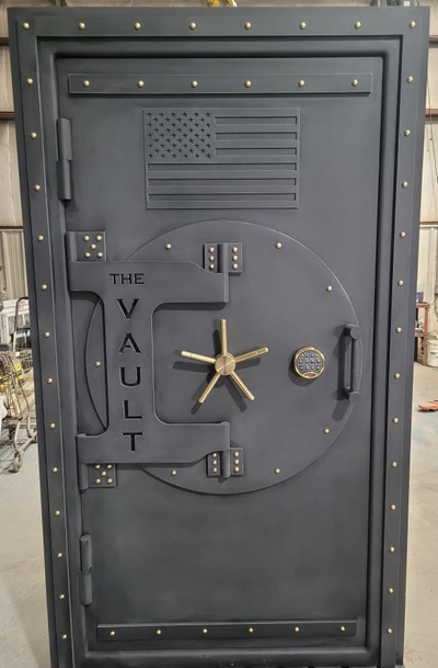 Large grey vault door with gold handle and fittings. It has an American flag engraved in the upper portion while the left side has the words 'The Vault' engraved into it.