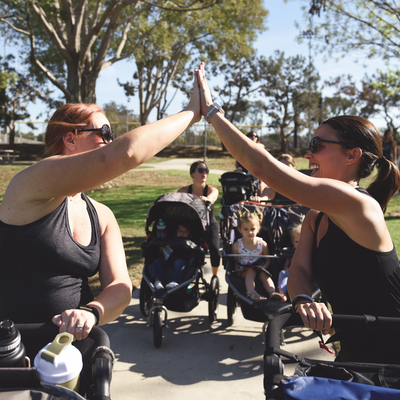 Moms giving high fives while pushing strollers in the park at a fit for mom stroller strides class.png