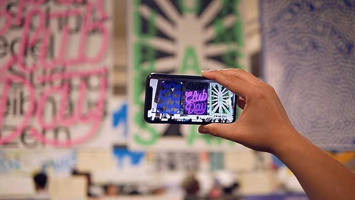 Hand holding a phone up to a wall of posters. The screen has AR writing on it 4-8