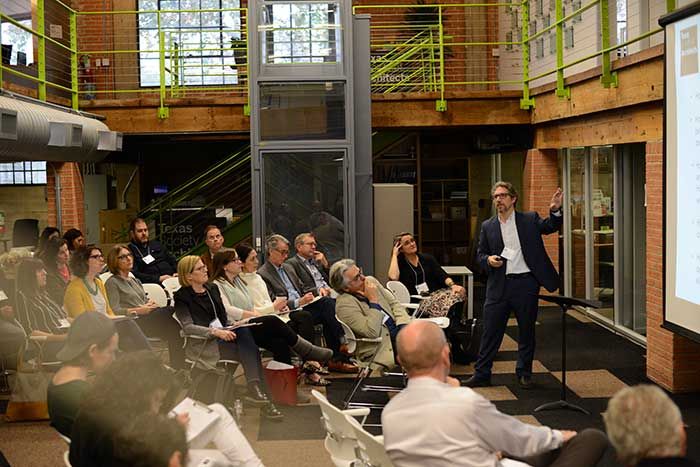 Man standing in front of a room pointing to a screen while a group of audience members watch 4
