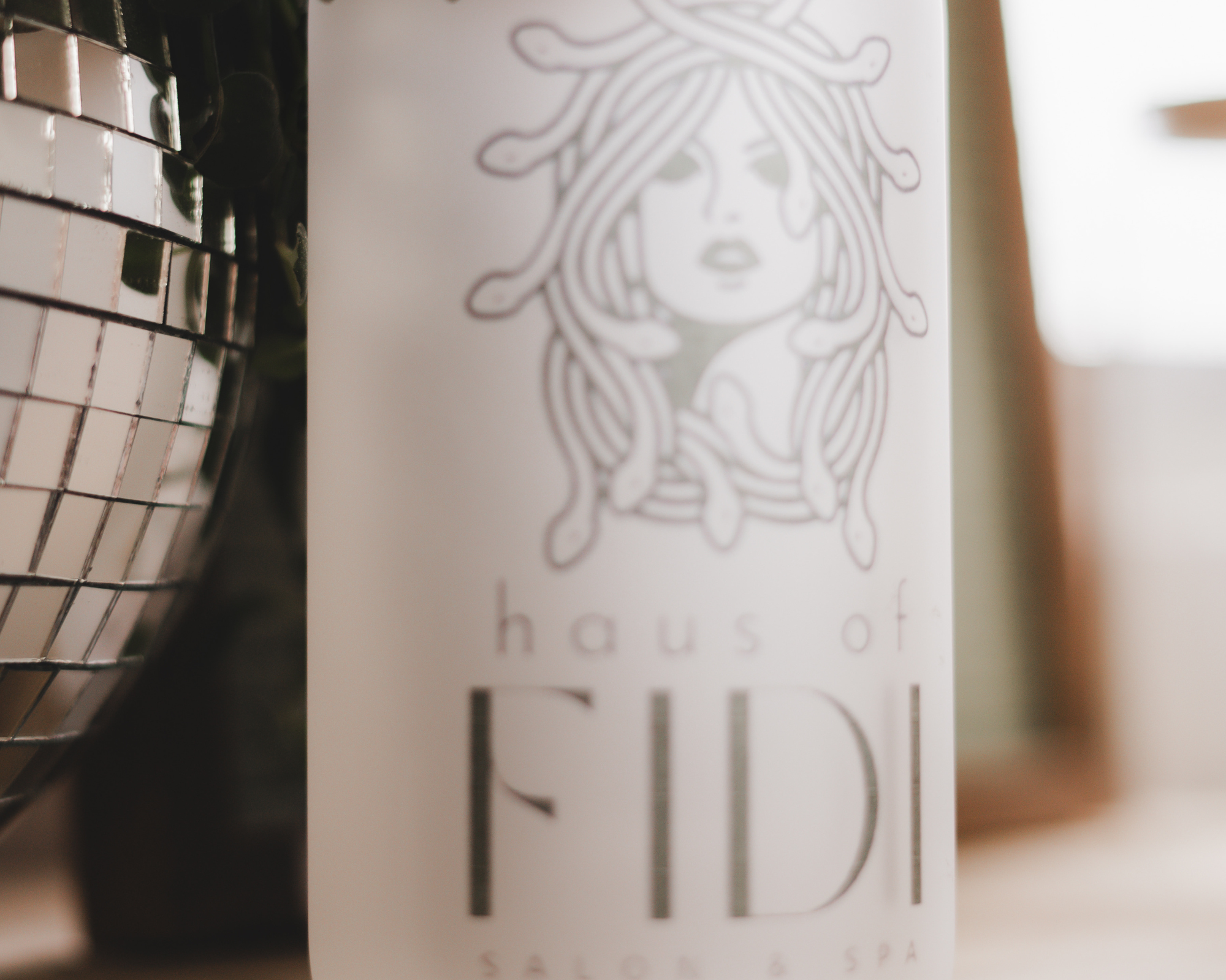 A Haus of FIDI logo-etched bottle beside a disco ball, representing the salon’s signature self-care products and modern branding in Pensacola, Florida. Haus of FIDI salon and spa product bottle