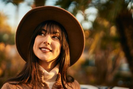 Woman with long brunette hair and warm caramel highlights wearing a brown wide-brim hat outdoors in autumn. Fall Hair Color Inspiration at Wild Hare Salon