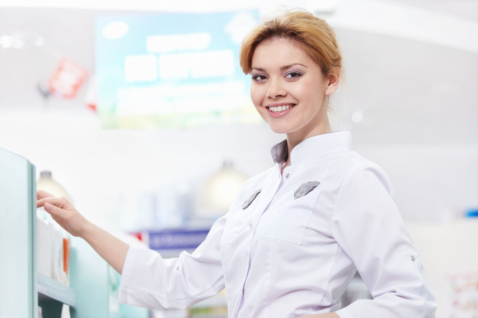 Blonde female pharmacist wearing a white coat smiles while standing near pharmacy shelves, symbolizing professionalism and customer service in a healthcare environment. Smiling Female Pharmacist in White Coat