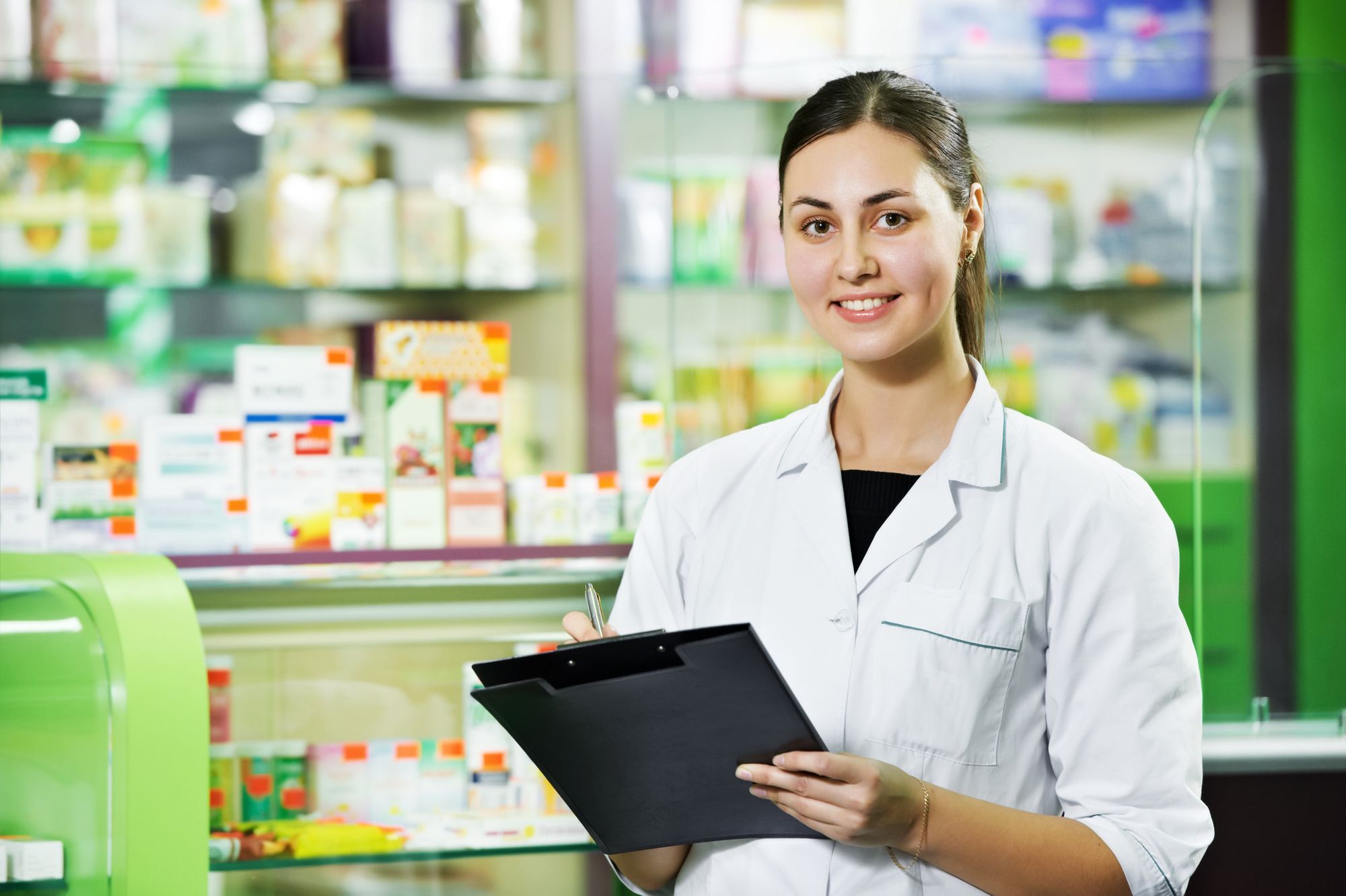 Young female pharmacist in a white coat smiling and holding a clipboard in front of pharmacy shelves, representing personalized support for compounded ivermectin. Pharmacist Holding Clipboard at Rock Ridge Pharmacy
