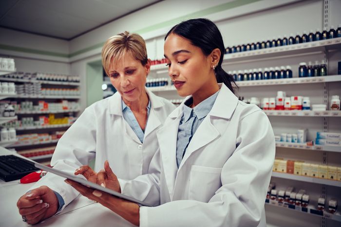 Two woman pharmacists in front of a wall of bottles and medication, and boxes looking at information Two woman pharmacies consulting